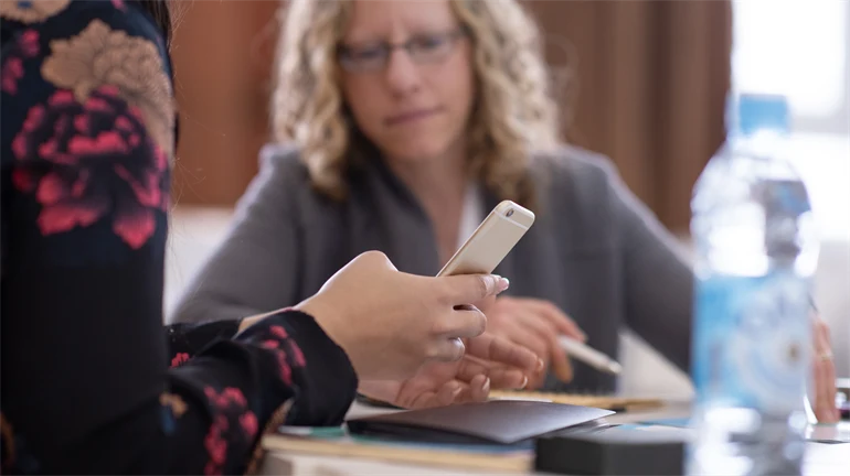 Kvinnlig hand vid ett bord som håller i en mobiltelefon, suddig kvinna i bakgrunden.