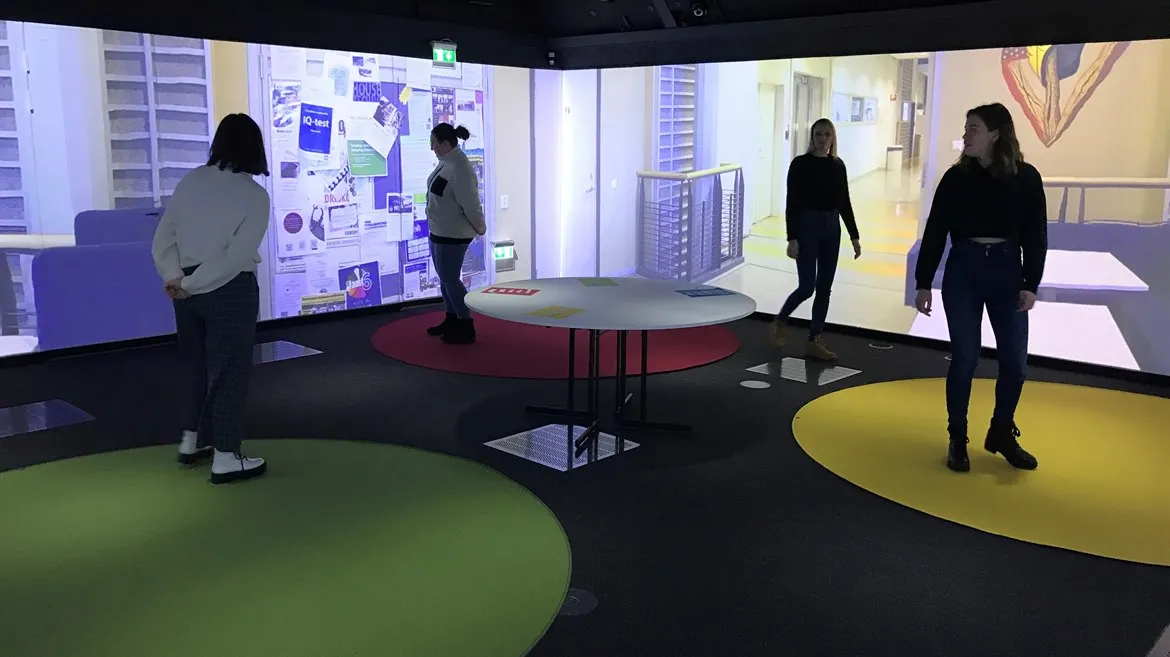 Fyra personer står i ett rum. På golvet finns runda mattor i olika färger. Några av personerna står på mattorna. På väggarna projiceras bilder från en korridor i en skolbyggnad.