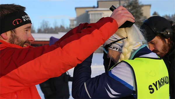 Man i röd jacka hjälper synskadad, kvinnlig skidåkare att bära skallbenshörlurar 