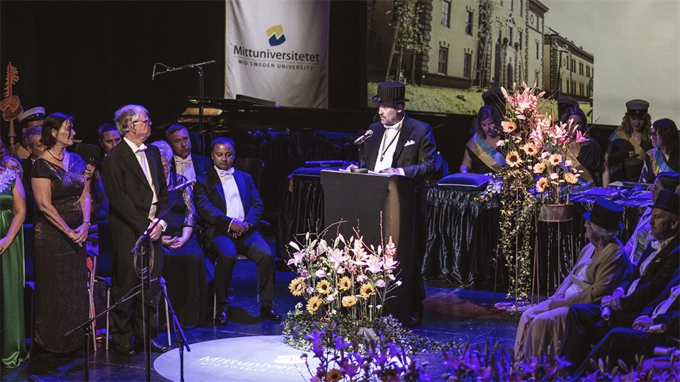 A ceremony stage with flower decorations, people, monitor, flags and a speaker.