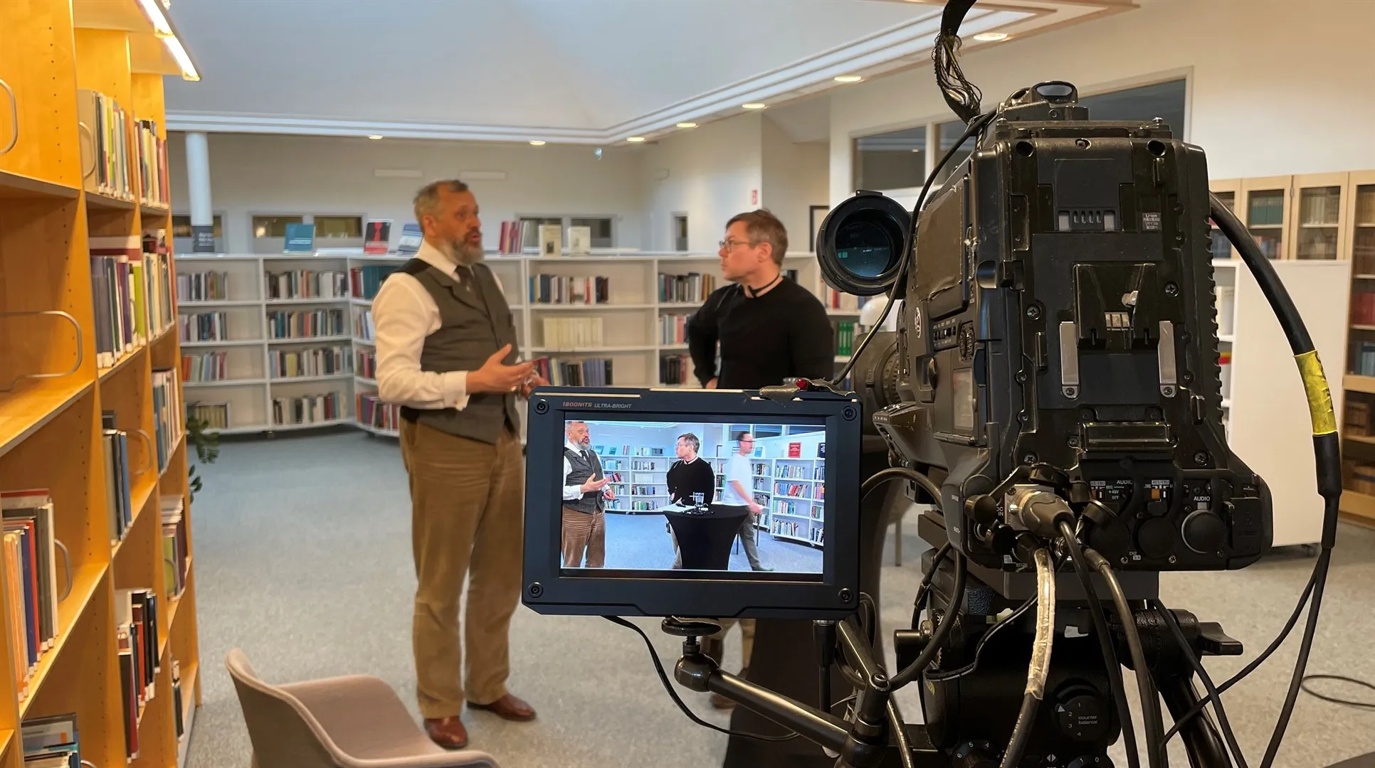 Två män står i ett bibliotek och pratar med varandra. I förgrunden syns en filmkamera. Männen syns även på kamerans skärm.