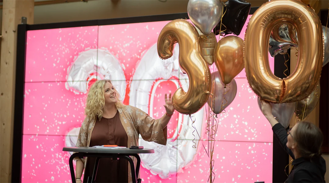 En kvinna står på en scen. Bredvid henne svävar ett antal ballonger i guld och silver. Två av ballongerna är formade som siffrorna 3 och 0. Hon tittar på en ballong och rör vid den.