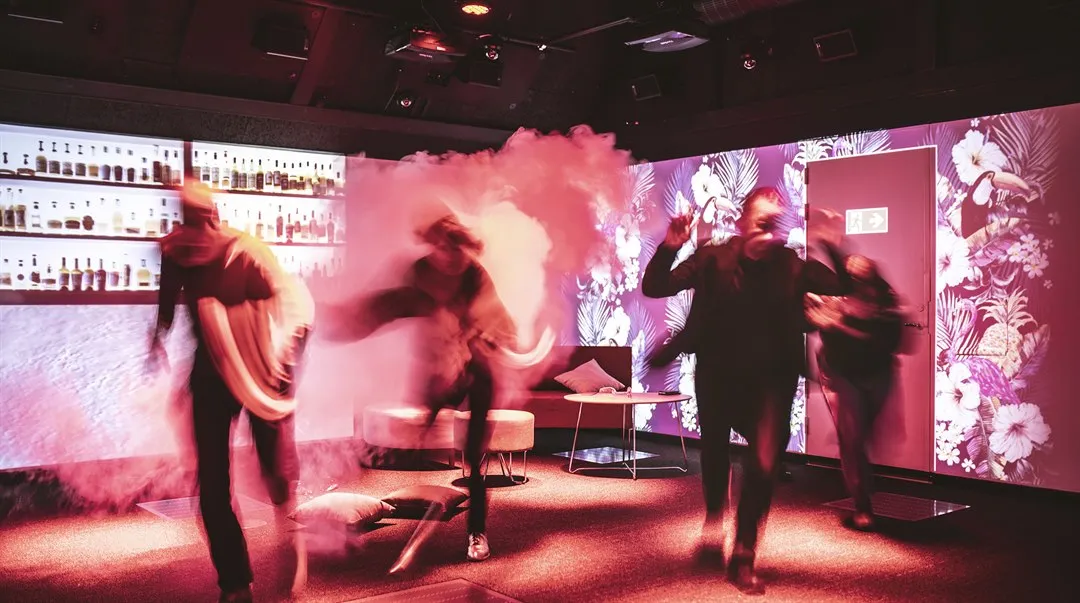 A room with smoke in the background. On the walls, a nightclub environment with a bar and an emergency exit is projected. In one corner are some poufs and a low table. Four people are running.